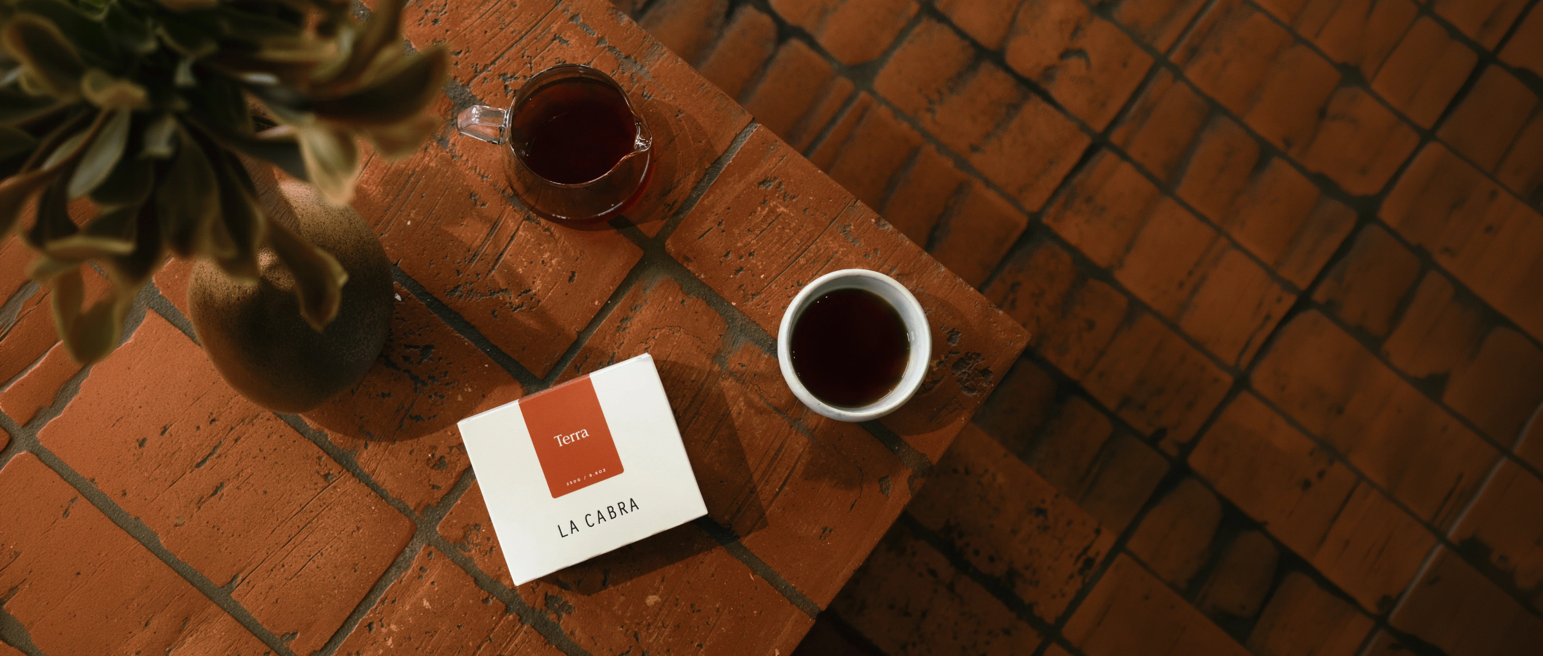 A styled overhead shot of a coffee table scene featuring a white box of La Chiara coffee beans, a white ceramic mug filled with dark coffee, a glass cup, and a potted plant on a reddish-brown brick surface.